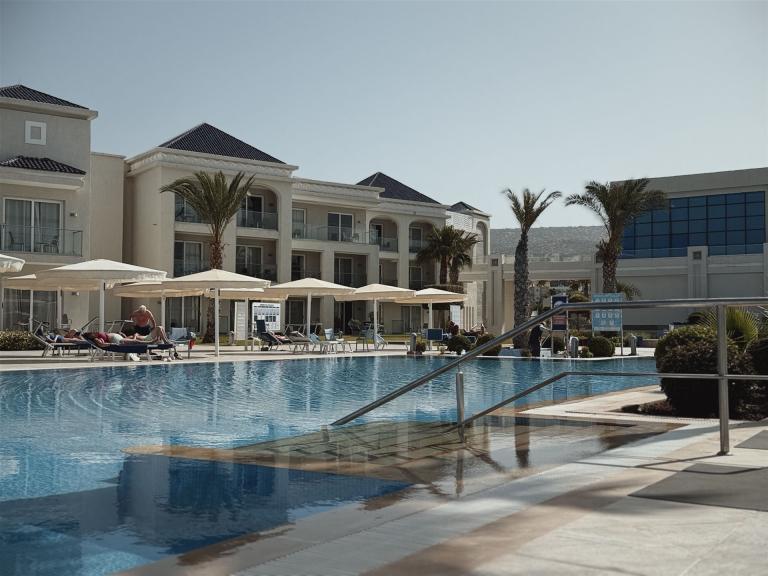 Ein großer Poolbereich eines modernen Resorts vor einem mehrstöckigen Hotelgebäude mit Palmen und Sonnenschirmen.