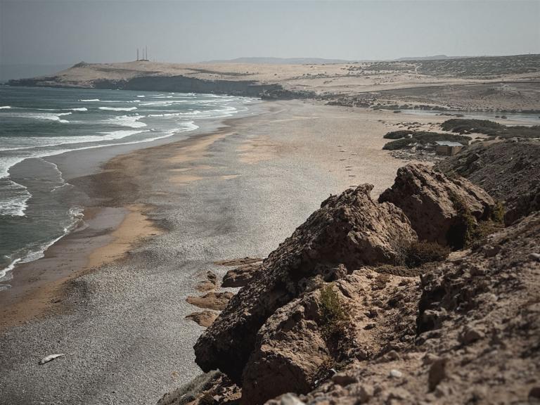 Küstenpanorama mit felsigem Vordergrund, einem breiten Sandstrand und ariden Hügeln im Hintergrund.