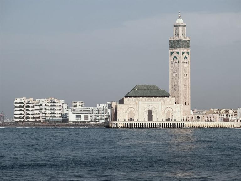 Eine große Moschee mit Minaretten auf einer Insel, umgeben von Wasser und modernen Hochhäusern im Hintergrund.
