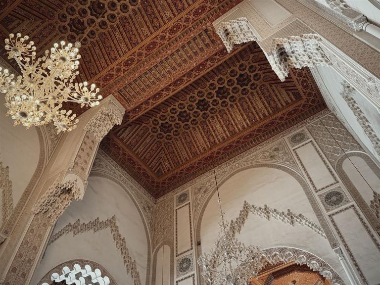 Innenansicht einer prunkvollen Halle mit reich gemustertem Deckenbaldachin und mehreren Kristalllüstern.