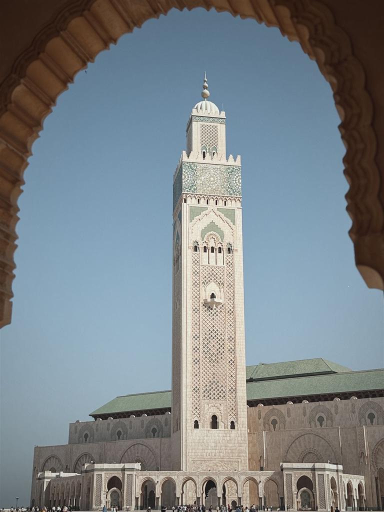 Ein hoher Minarett im islamischen Baustil vor einem großen Gebäude, eingerahmt von einem bogenförmigen Durchgang.