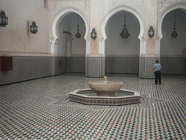 Ein großer Innenhof mit geometrischem Mosaikboden, umgeben von gewölbten Arkaden und einem runden Wasserbecken.