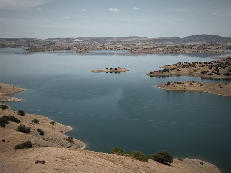 Eine weite Landschaftsaufnahme eines Sees, umgeben von trockenen, hügeligen Landschaften mit einer kleinen Insel im Zentrum.